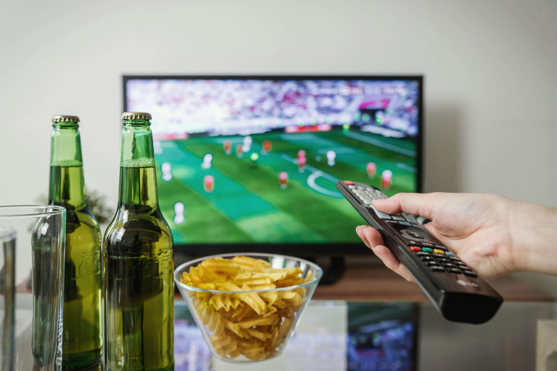 Hand holding remote control with TV displaying soccer match, two beer bottles, and bowl of chips on table