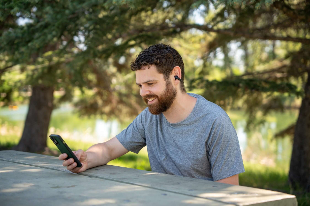 Man enjoying music on his phone while wearing Gibutech Ultra bone conduction earbuds at a park.